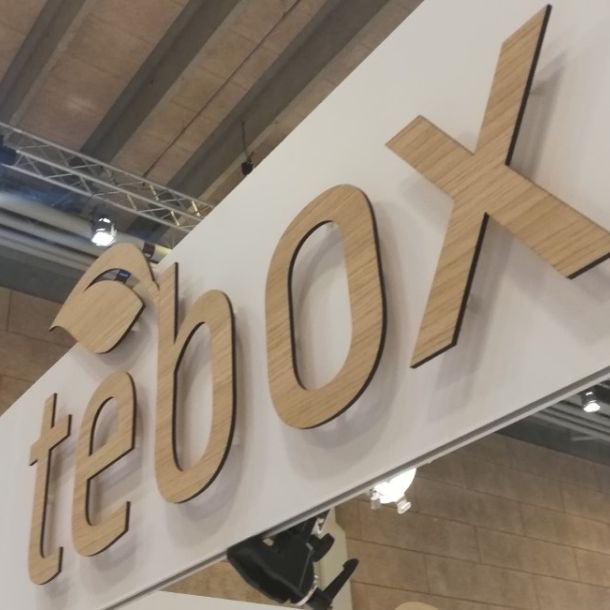 Stort Tebox logo i træ monteret på hvid bagplade på messestand.