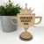 Pokal i træ med teksten Verdens Bedste Far. Står på en hylde med en grøn plante.