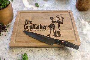 Skærebræt på bord med urter. På skærebrættet er graveret teksten: The Grillfather og der er en mand der står ed en grill.