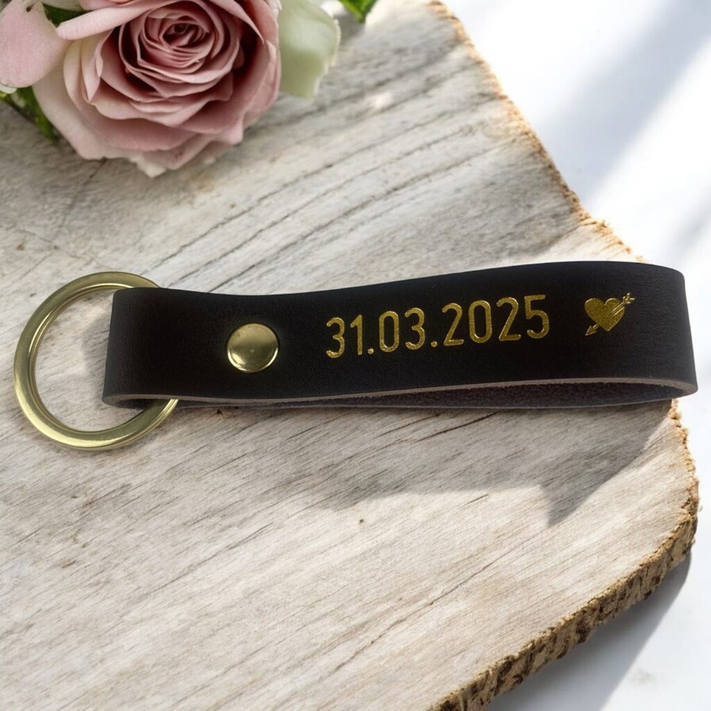 Brun lædernøglering med guld tryk. Der er trykt en data som markerer kærlighed. Ligger på et rustikt bord med en rose.