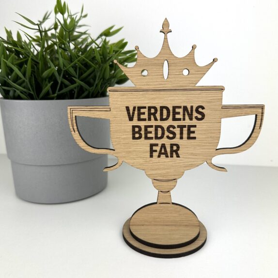 Pokal i træ med teksten Verdens Bedste Far. Står på en hylde med en grøn plante.